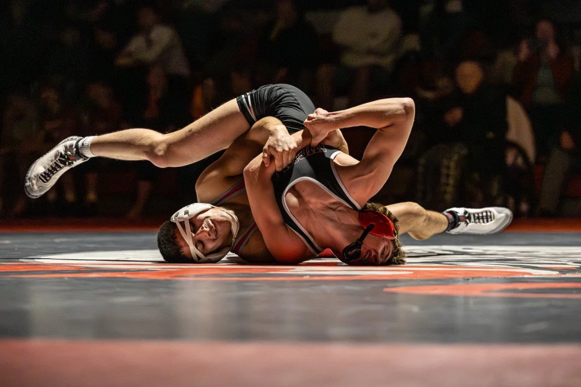 Two wrestlers compete on a wrestling mat, with one wrestler on the ground attempting to hold the other in a wrestling move, both wearing wrestling singlets and headgear.