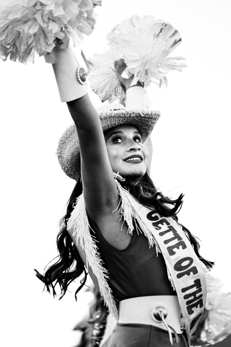 A woman dressed as a cheerleader, wearing a sash that reads "COSTE OF THE", a wide-brimmed straw hat, and holding pom-poms, cheering with her arm raised.