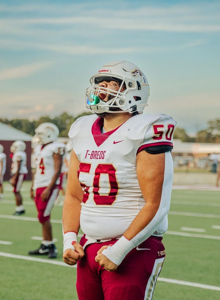 A football player in a white and red uniform with the number 50 and the team name T-BREDS on the jersey, wearing a white helmet with a face guard, standing on a football field, celebrating with clenched fists.
