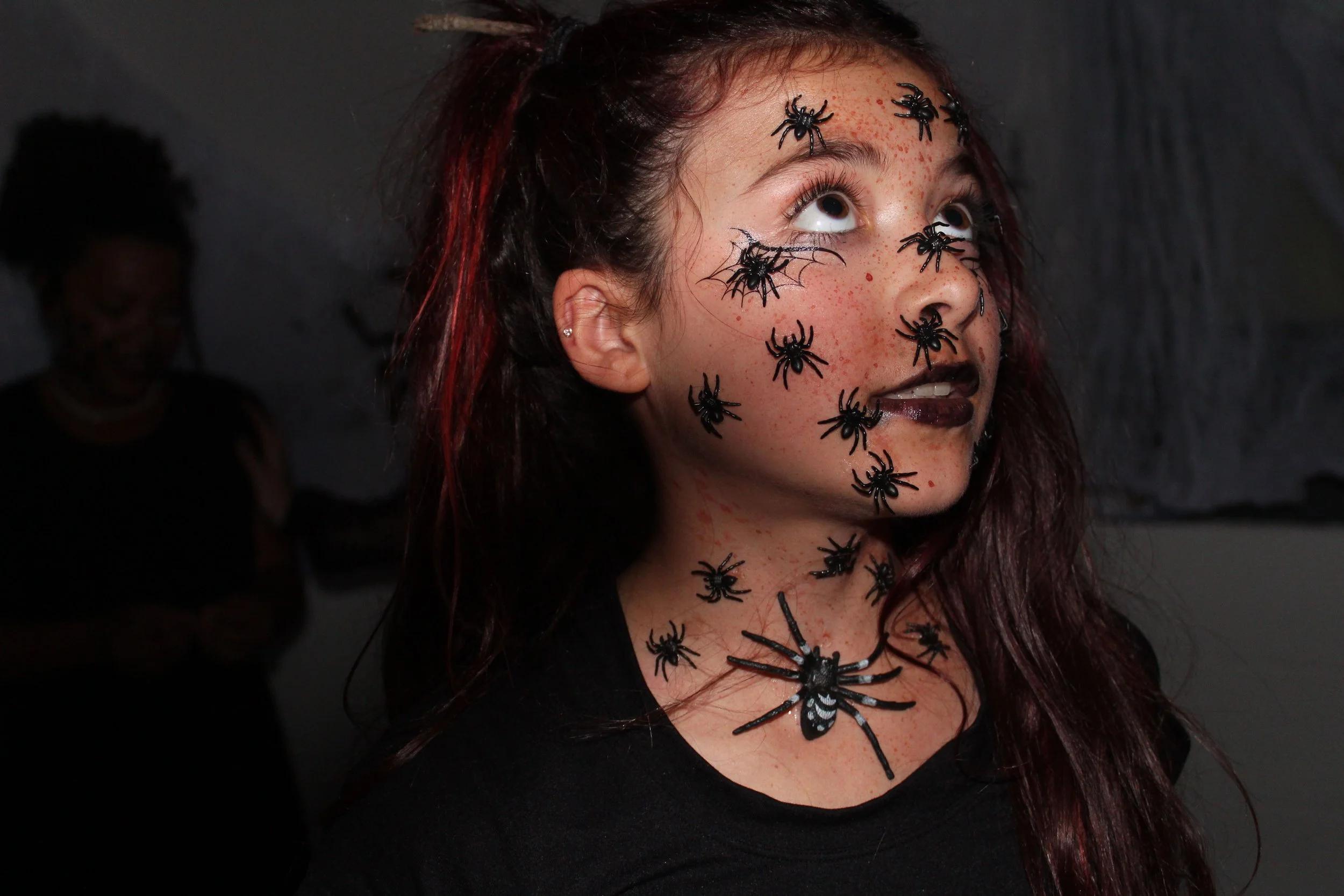 A woman with dark red hair and freckles is decorated with small black plastic spiders and a black butterfly on her face and neck, dressed for Halloween.
