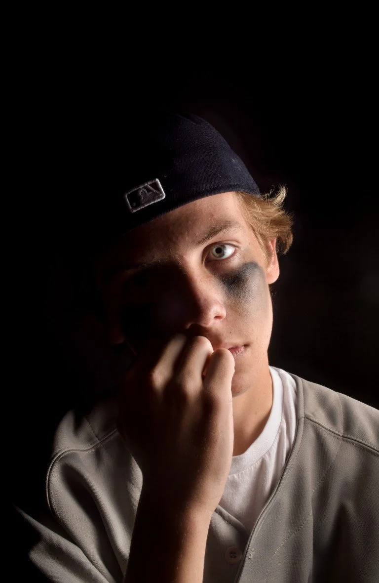 A young man with a black eye makeup, wearing a black baseball cap and a light-colored shirt, holding his hand near his face in a dark setting.
