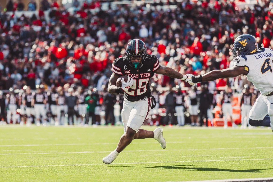 A college football game with a Texas Tech player running with the ball while a West Virginia defender tries to tackle him on a grassy field crowded with spectators.