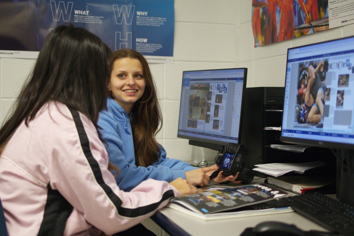 Two girls sit at a desk in a classroom, looking at each other and smiling. One girl has long dark hair and is wearing a pink jacket, while the other girl has long brown hair and is wearing a blue hoodie. There are two computer monitors on the desk di