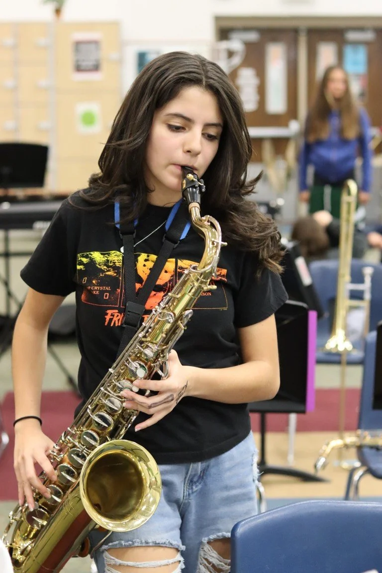A young woman with long brown hair playing a gold alto saxophone in a music classroom, with other students and musical instruments in the background.