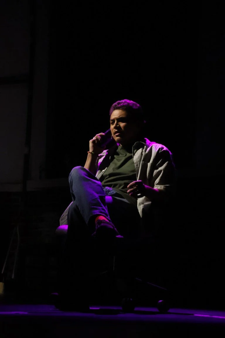 Woman sitting on a chair in a dark room, illuminated by purple and pink stage lighting, talking on a cellphone.