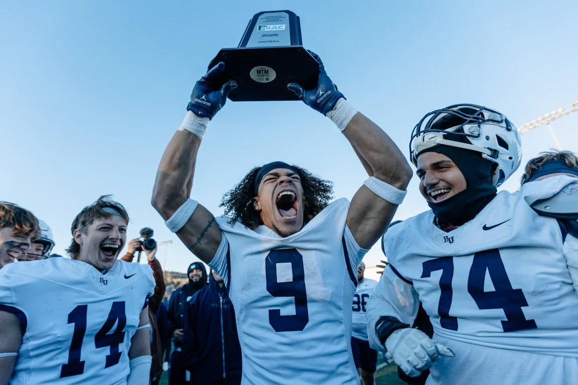 A group of football players celebrating, with one player lifting a trophy overhead, wearing a white jersey with the number 9, surrounded by teammates, smiling and cheering.