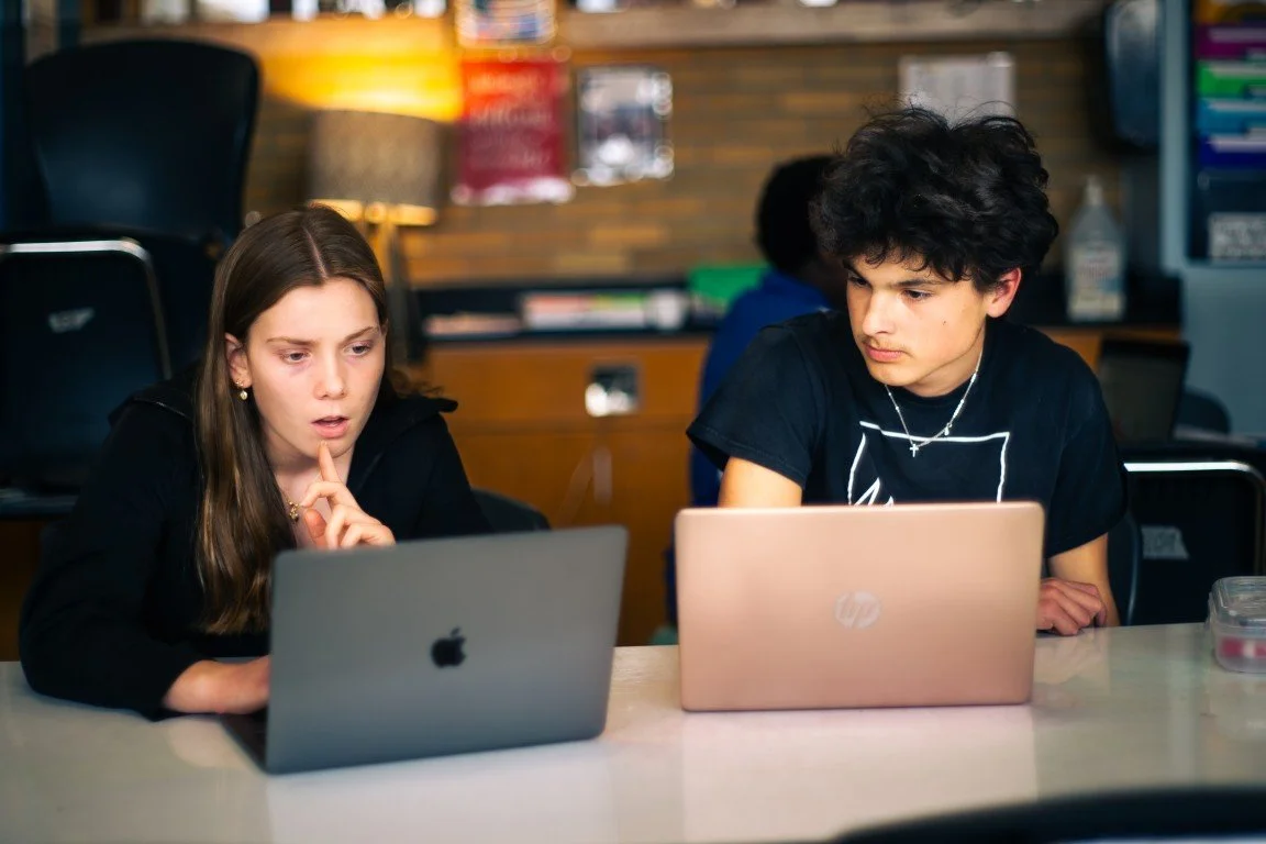Two teenagers, a girl and a boy, sitting at a table in a classroom or study area, focused on their laptops. The girl appears puzzled, while the boy is looking at his screen. There are chairs, a lamp, and shelves in the background.