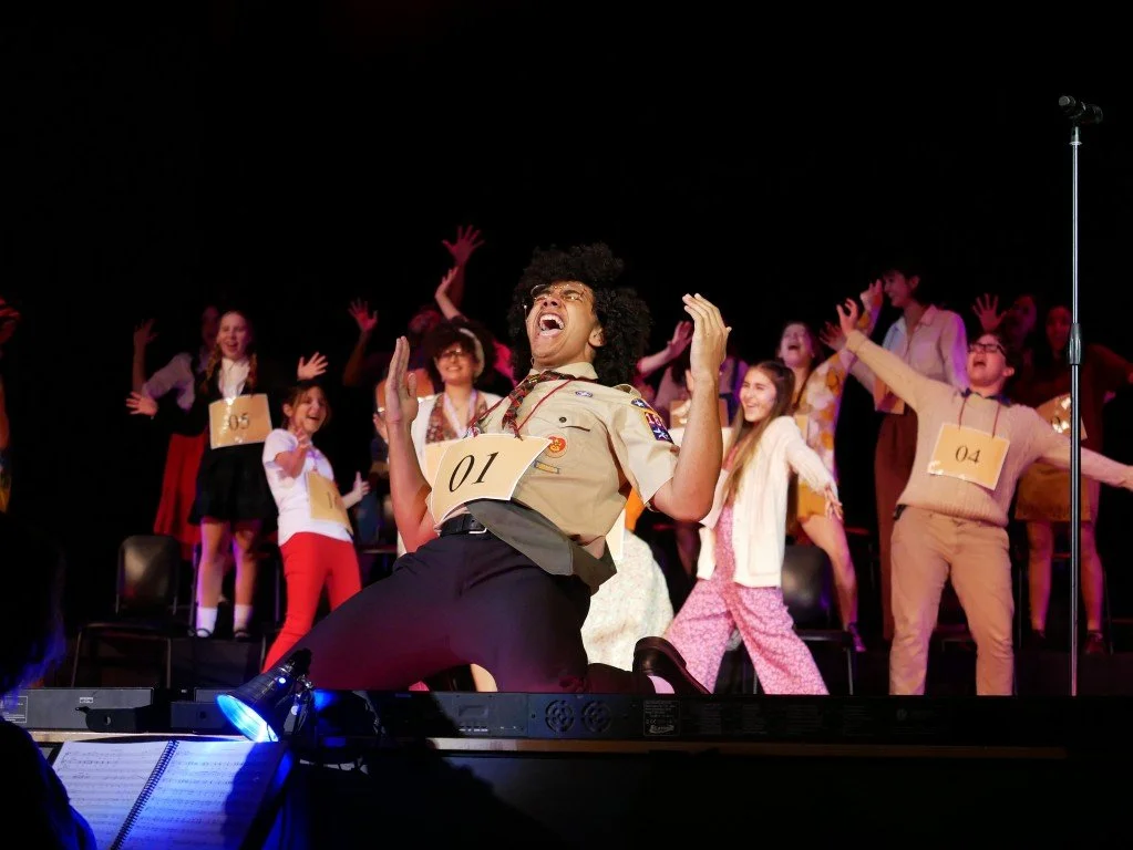 A person in a scout uniform dancing on stage with children, some with numbered paper tags, in a theatrical performance setting.