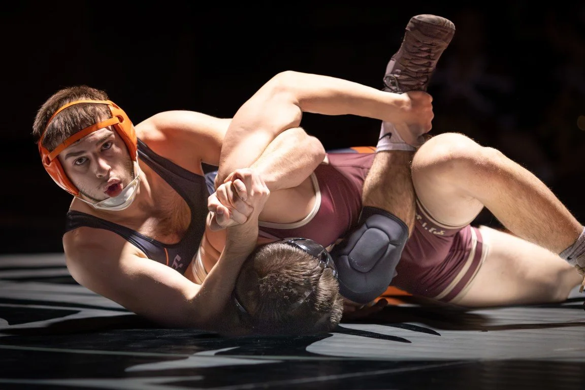 Two male wrestlers in a match, one on top of the other, wrestling on a mat with black and white patterns, one wearing an orange protective headgear and the other in black headgear.