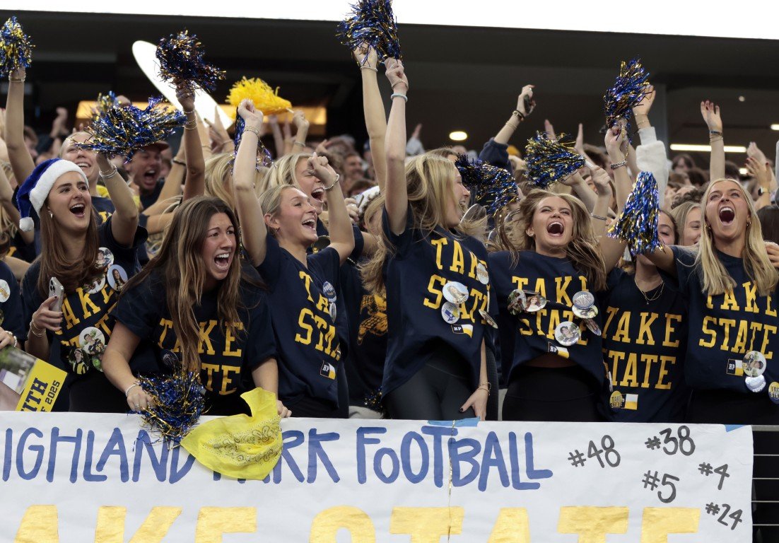 Group of enthusiastic young women cheering at a Highland Park football game, wearing team shirts and holding pom-poms, with a banner displaying football player numbers.