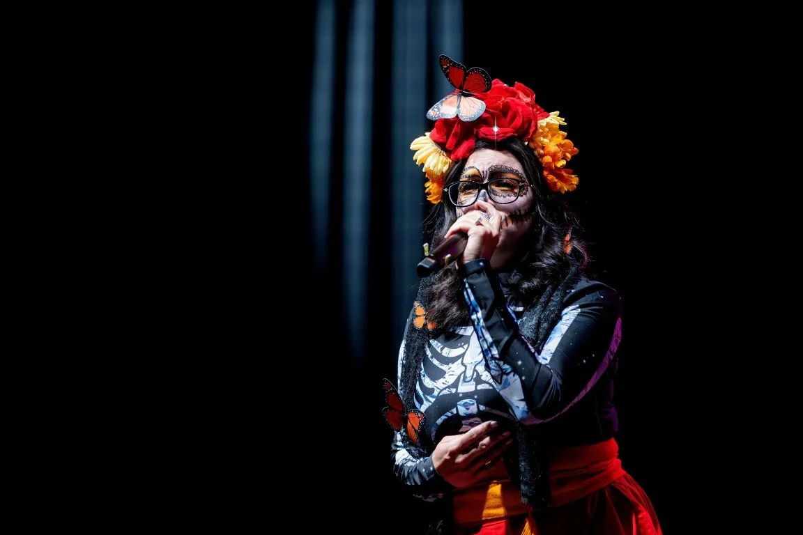 A woman wearing a flower crown and glasses, holding a microphone and singing on stage. She has face makeup resembling a Day of the Dead skull and butterflies on her outfit.