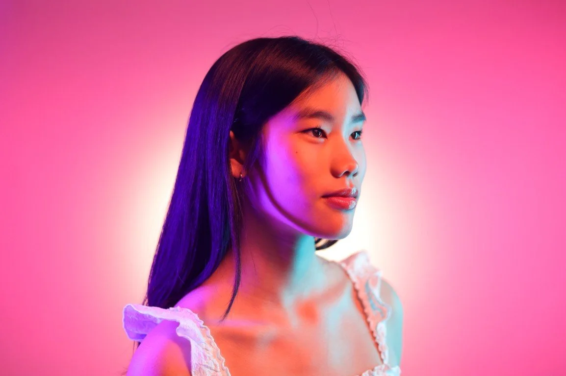 Portrait of a young woman with long dark hair against pink and purple lighting.