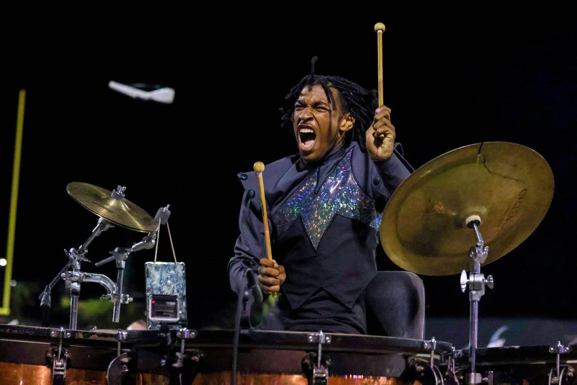 A drummer passionately playing a drum set with a facial expression of intense emotion, holding drumsticks in mid-motion, against a dark background.