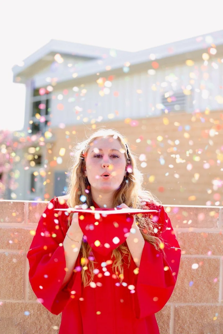 Young woman in a red graduation gown blowing confetti in front of a brick wall and house.