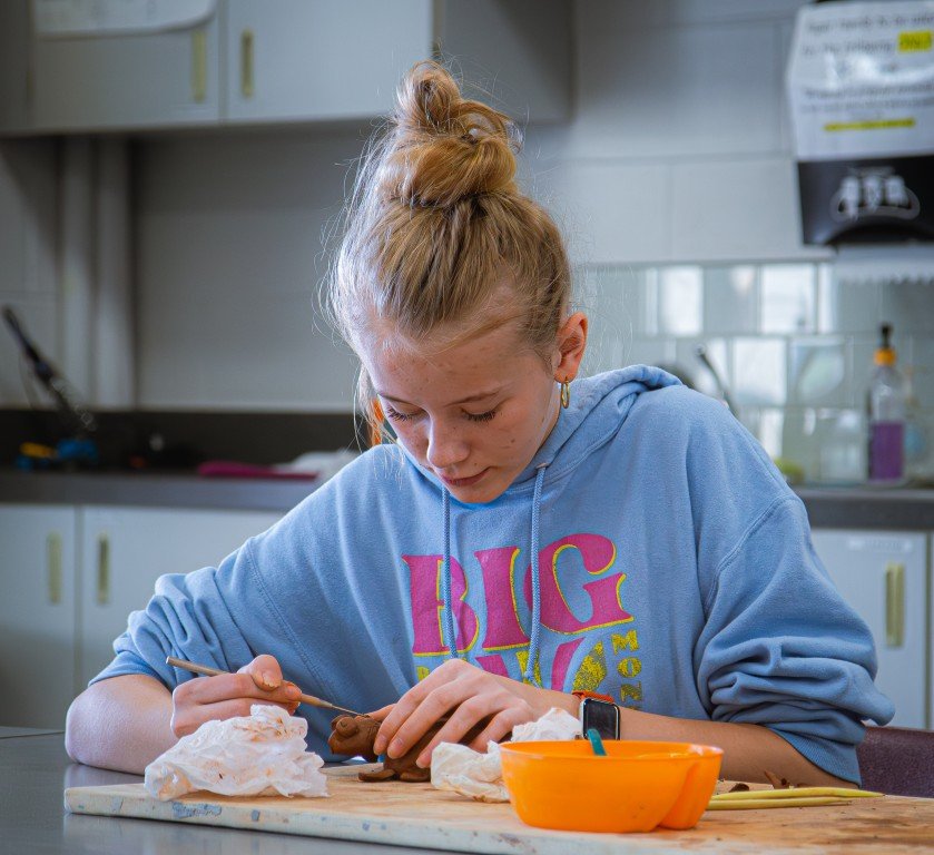 A girl with red hair in a bun, wearing a blue hoodie, working on a clay project at a table in a classroom.