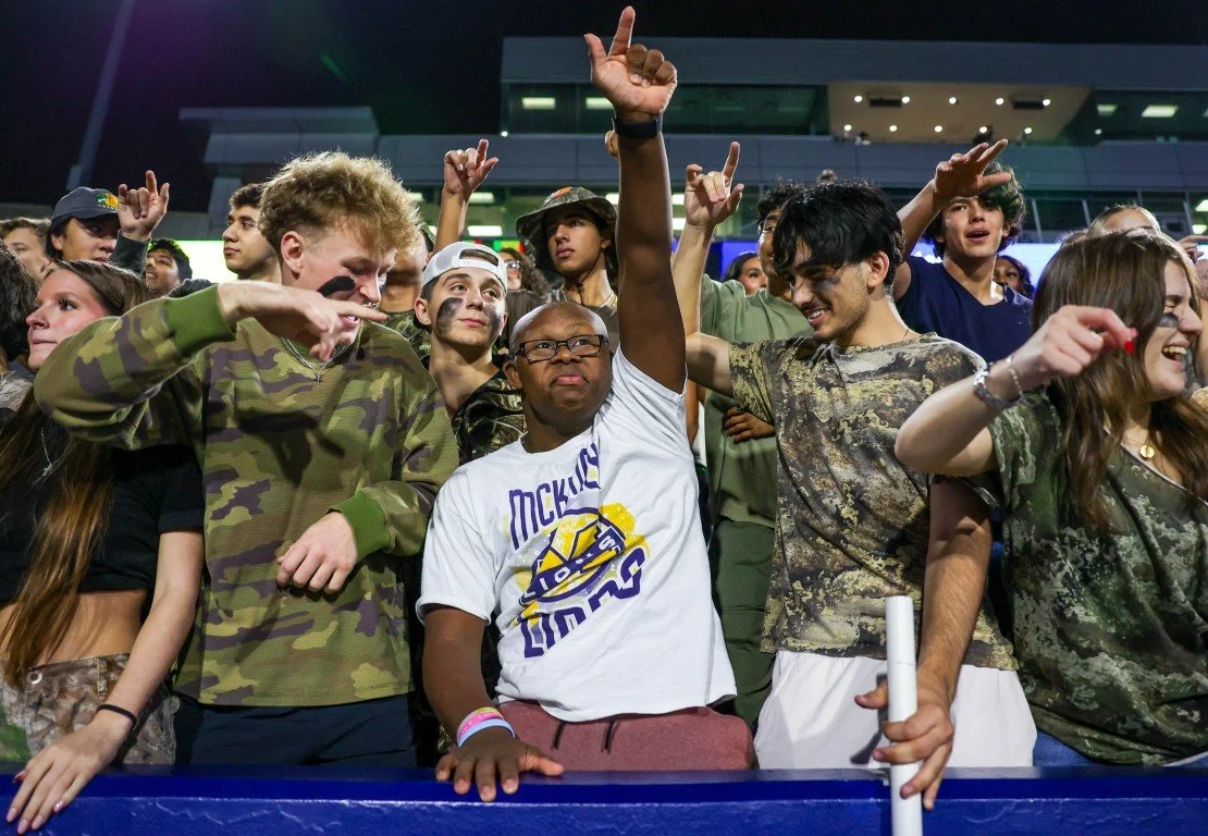 Crowd of young people at a concert or event, many dressed in camouflage, some raising hands, some smiling, in an outdoor setting at night.