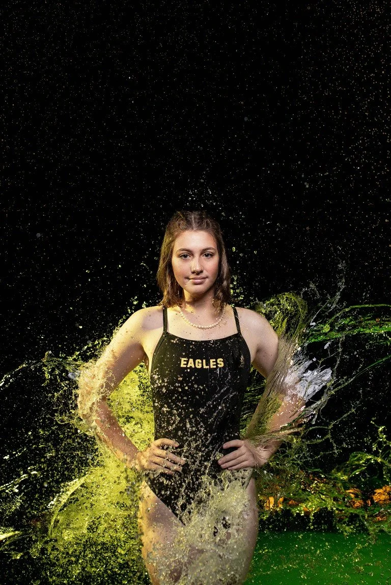 Young woman in a black swimsuit with 'EAGLES' written on it, standing with hands on hips, surrounded by splashing water, against a dark background.