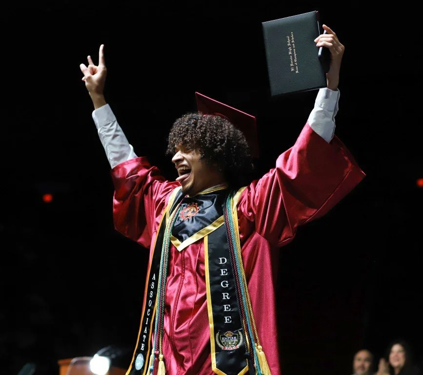 A young woman in a red graduation gown and cap celebrating on stage, holding a diploma and raising her hand in a peace sign.