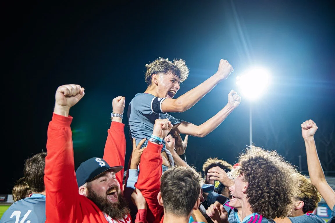 A group of joyful sports team members celebrating, lifting a young man on their shoulders under bright stadium lights.