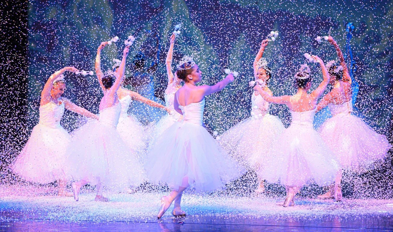 Six ballet dancers in white tutus performing on stage with falling confetti, illuminated by colorful stage lighting.