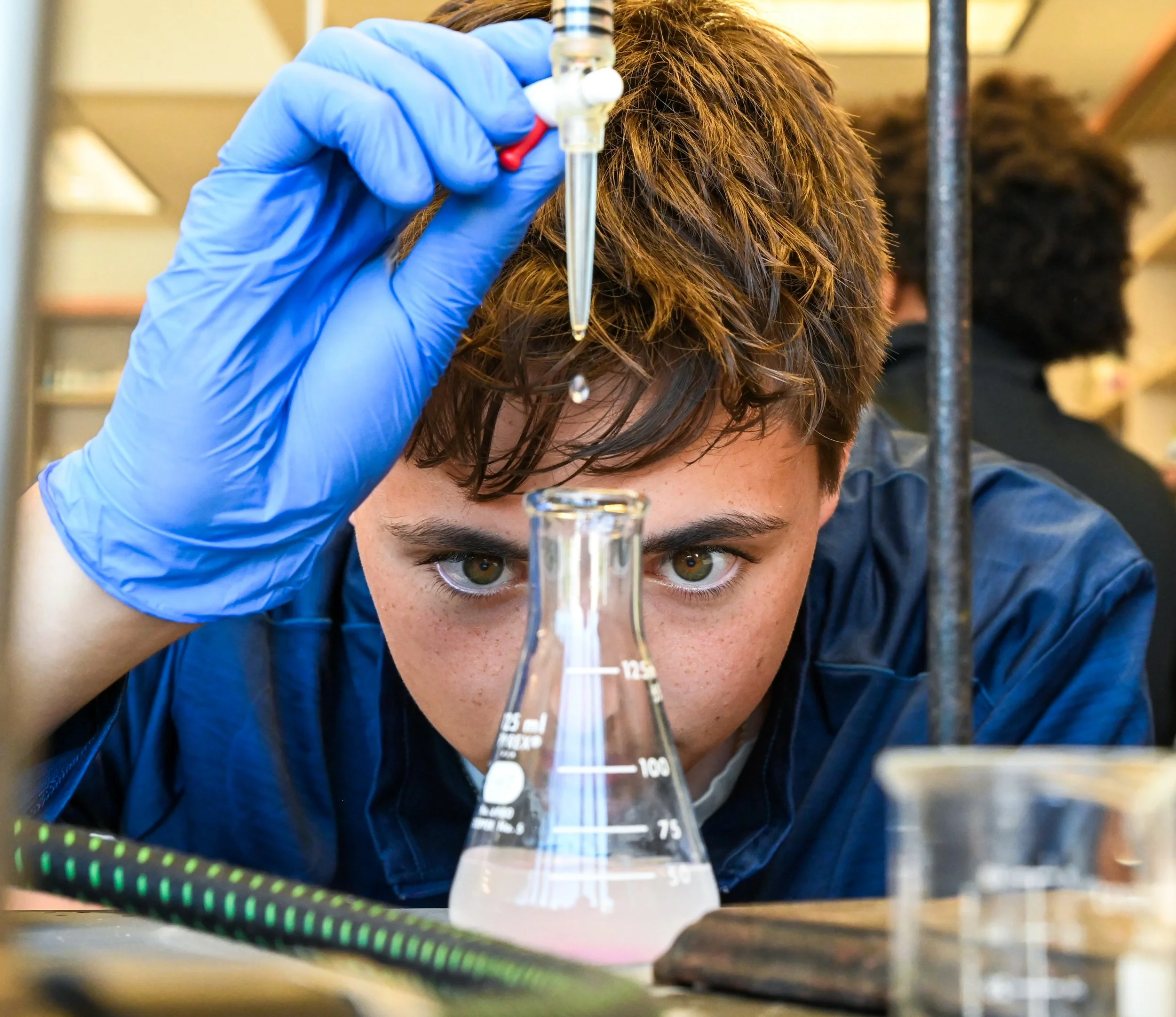 A person wearing blue gloves is closely examining or working with laboratory equipment, including a glass beaker and a pipette, in a science laboratory setting. The person's face is partially visible, with focus on their eye and hair.