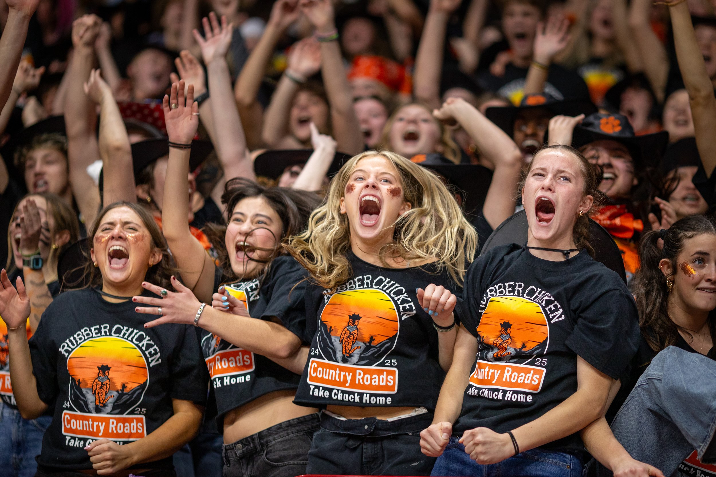 A large group of people at a rally or celebration, with many raising their hands and cheering. Several women in the foreground are wearing black T-shirts with a sunset and tree logo, and text that reads 'Rubber Chicken' and 'Country Roads,' indicatin