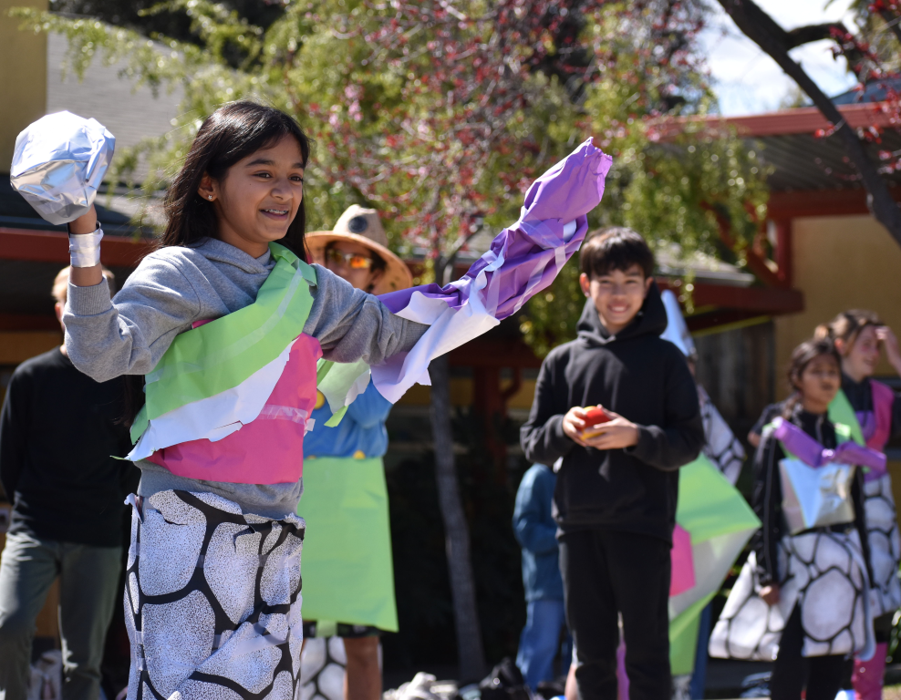 Smiling girl in colorful outfit with paper arms and white gloves, celebrating outdoors with other children.