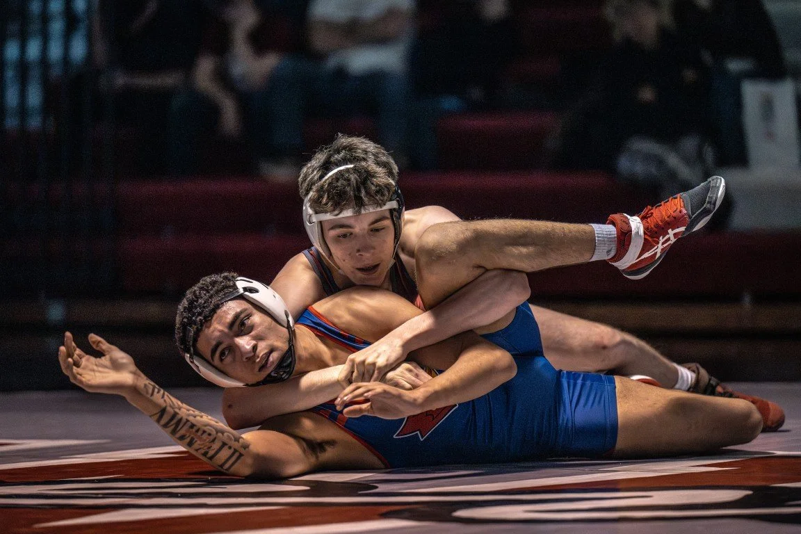 Two male wrestlers in singlets and headgear competing on a wrestling mat, with one on top of the other in an intense position.