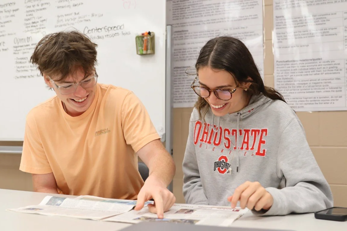 Two teenagers sitting at a table, smiling and looking at newspapers, in a classroom setting with notes and posters on the whiteboard behind them.