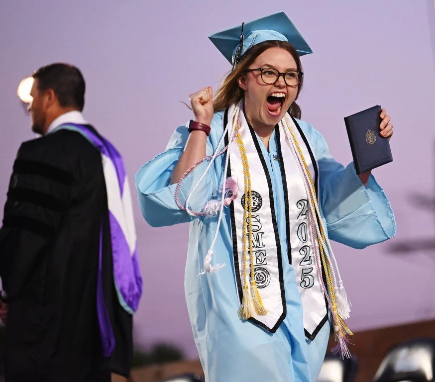 A woman in a light blue graduation gown and cap celebrating on stage, holding a diploma, with a broad smile and fist raised in excitement.
