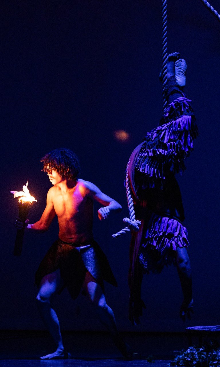 A shirtless man with curly hair holding a flaming torch, performing a dance on stage with dark background.