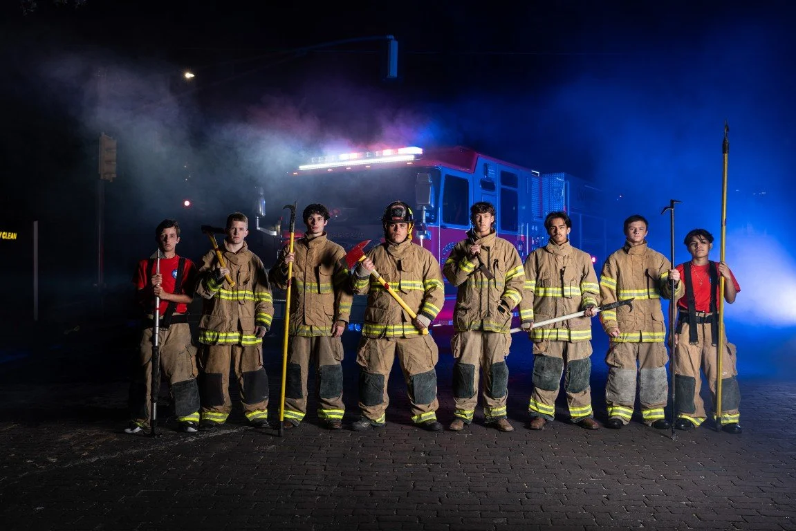 Group of firefighters standing in front of a fire truck at night, holding firefighting tools, with smoke and blue lights in the background.