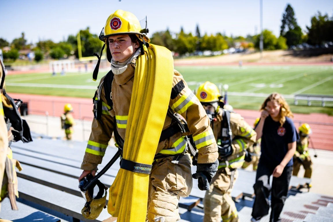 Firefighters in full gear, including helmets and oxygen tanks, walking on bleachers at an outdoor sports field.