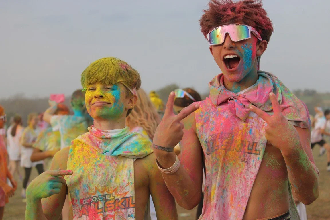 Two boys covered in colorful powder at a color festival, smiling and making peace signs.