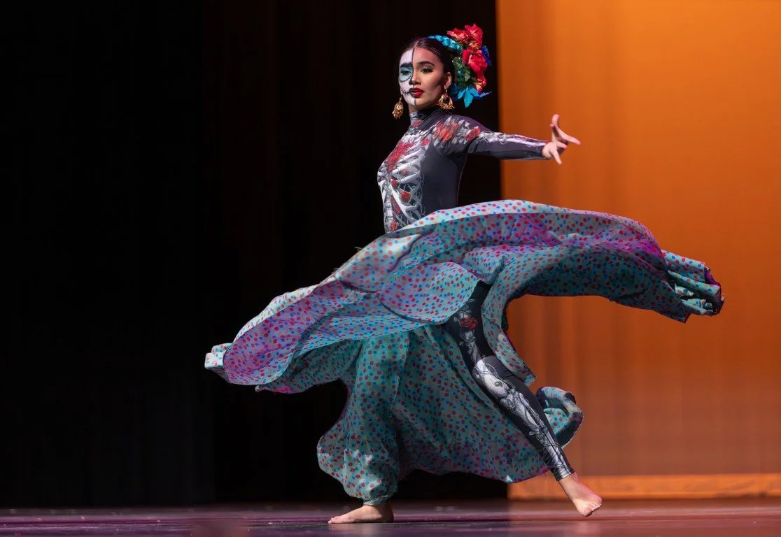 A woman in vibrant traditional costume dancing on stage with multicolored skirt and floral headpiece.