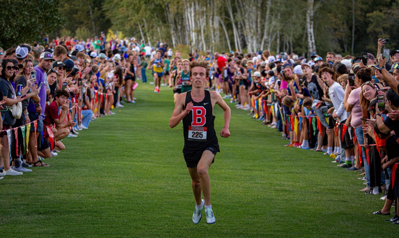 A male runner wearing a black tank top with a red letter B, black shorts, and white shoes running in a race, surrounded by a large crowd of spectators on both sides of the grassy track, many taking photos or videos.