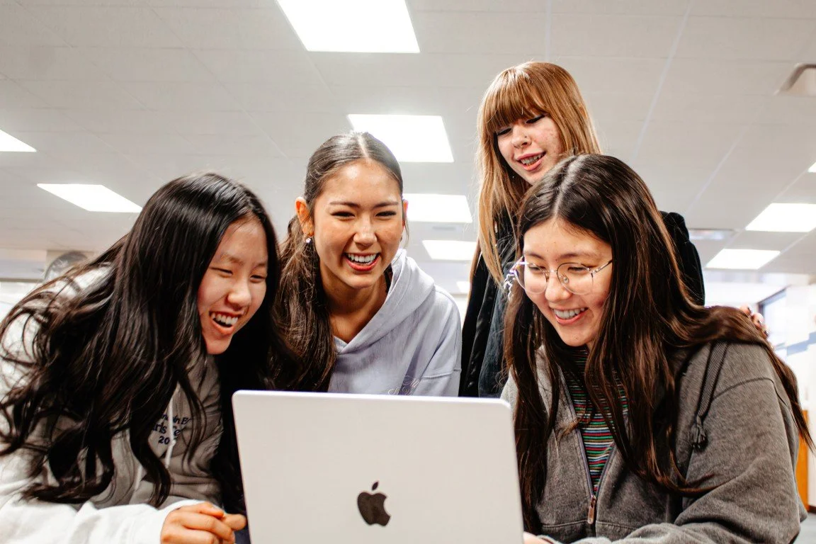 Four young women gathered around a laptop, smiling and laughing together.