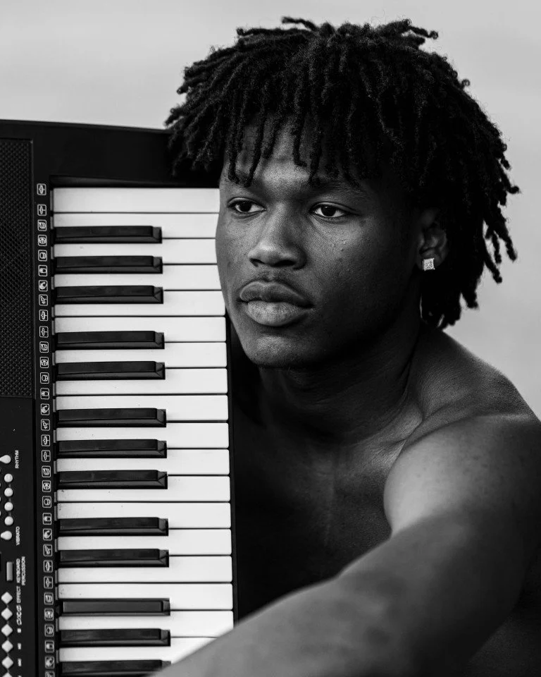 A young man with dreadlocks, shirtless, posing next to a keyboard with black and white keys, black background, in black and white photography.