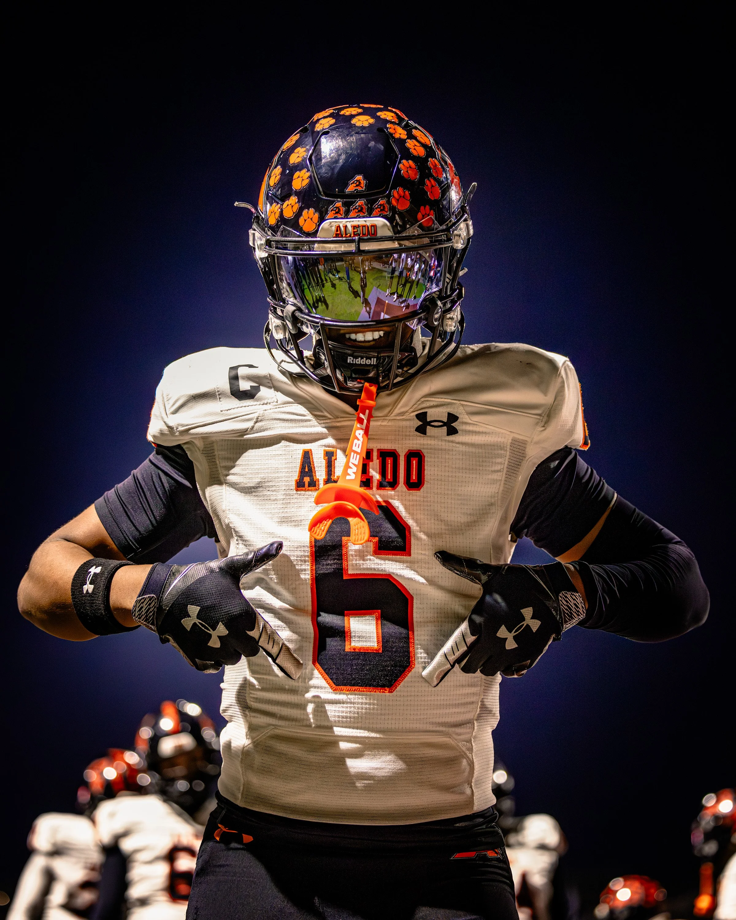 A football player in a white uniform with orange accents and black gloves, wearing a black helmet decorated with orange paw prints and the name Aledo, stands on the field at night with his hands on his hips, with teammates and stadium lights in the b