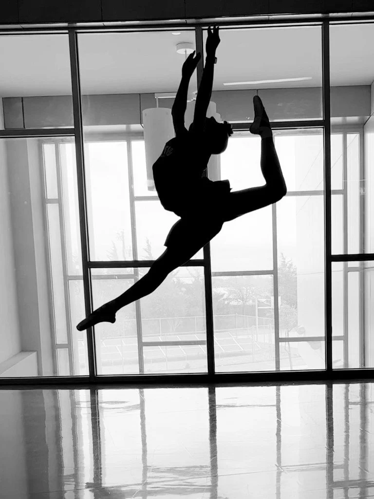 Silhouette of a person performing an acrobatic flip or dance move in front of a large window with natural light