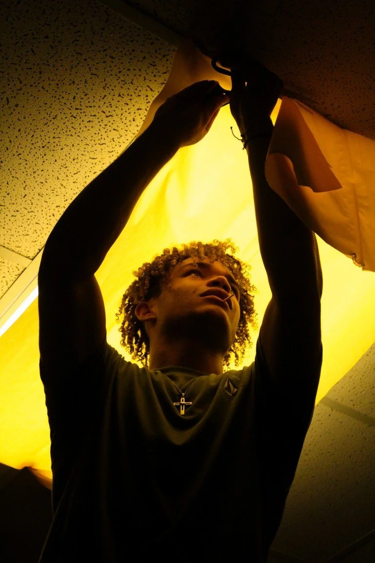A person with curly hair hanging ceiling tiles as they work, illuminated by a yellowish light from a suspended light fixture.