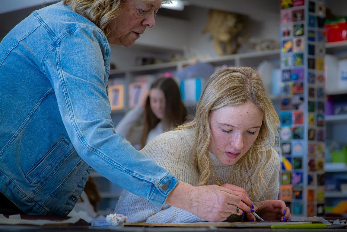 A woman with long blonde hair sitting at a table, working on an art project with a young girl, in a craft store aisle with colorful shelves.