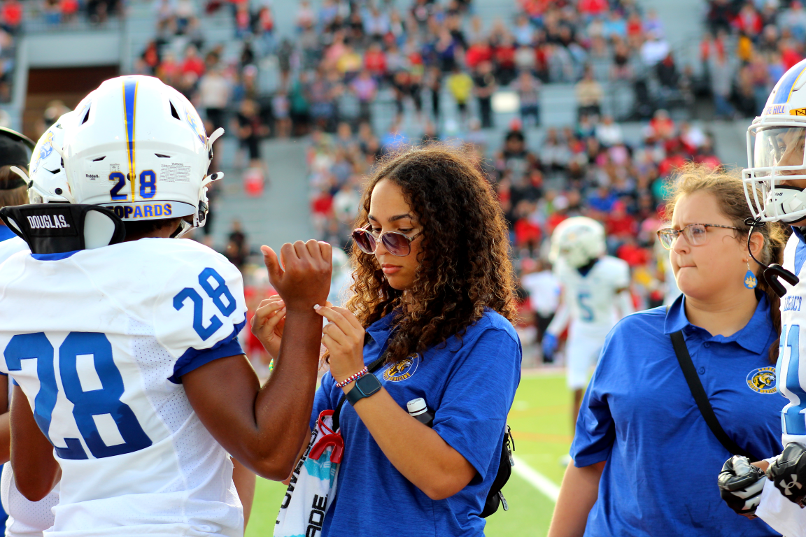 A woman in sunglasses and a blue shirt with a sports team logo is applying a medical tape or bandage to a football player wearing a white jersey with the number 28, at a football game with spectators in the stands.
