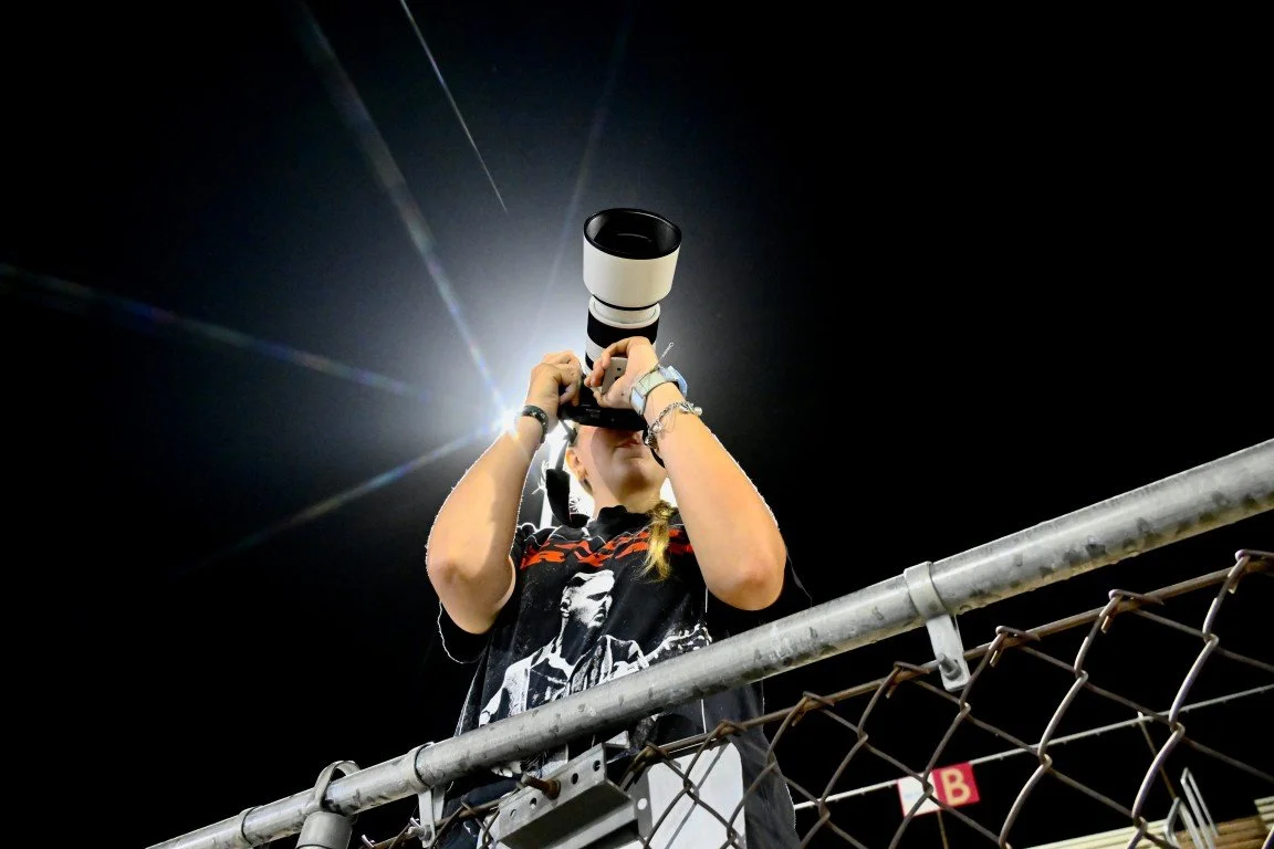 A woman with bracelets on her arms holding a camera with a large telephoto lens, taking a picture at night behind a chain-link fence.