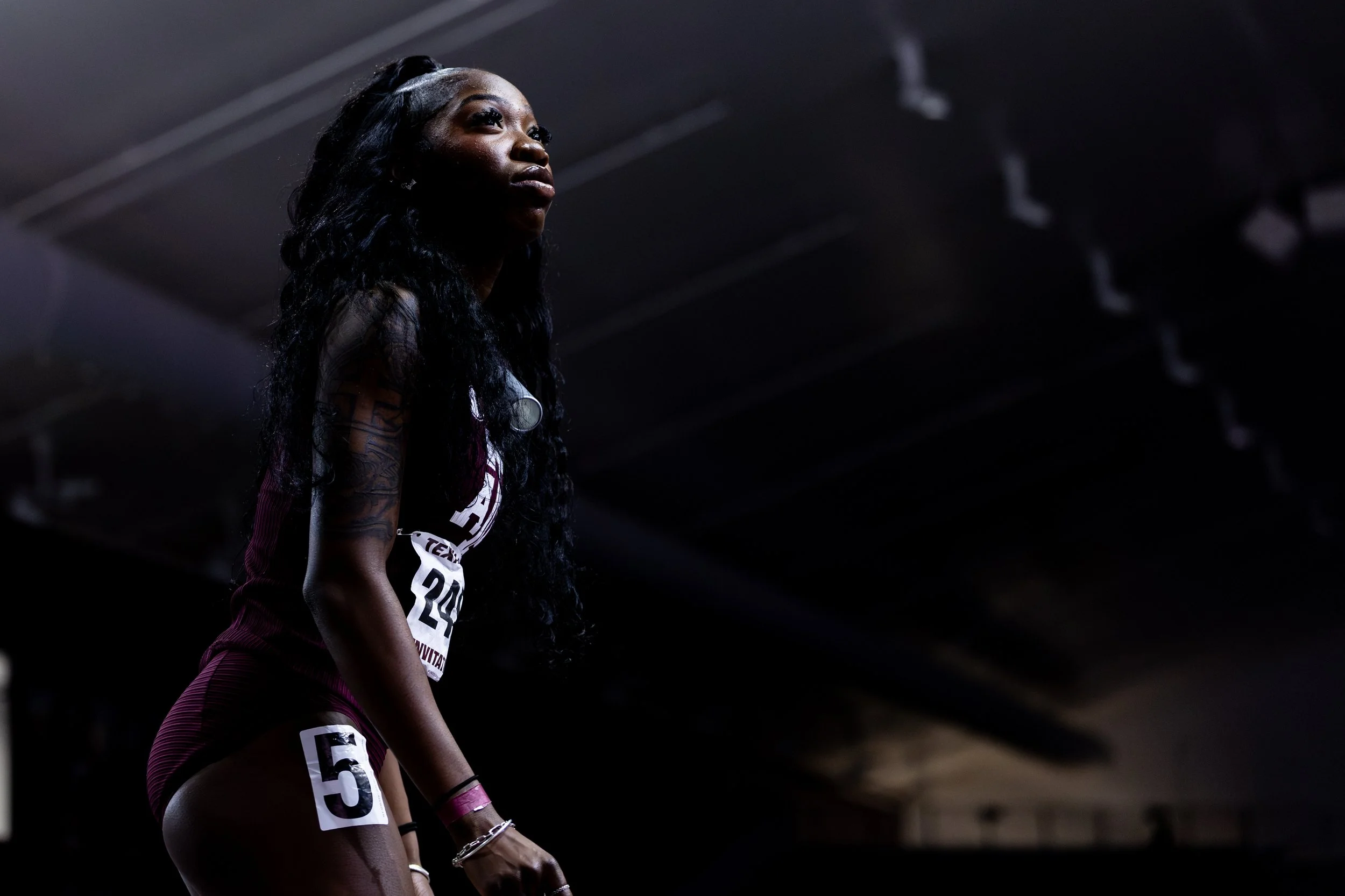 Female athlete wearing a maroon sports uniform with a number 5 on her shorts, standing in a dimly lit indoor sports arena, looking upward.