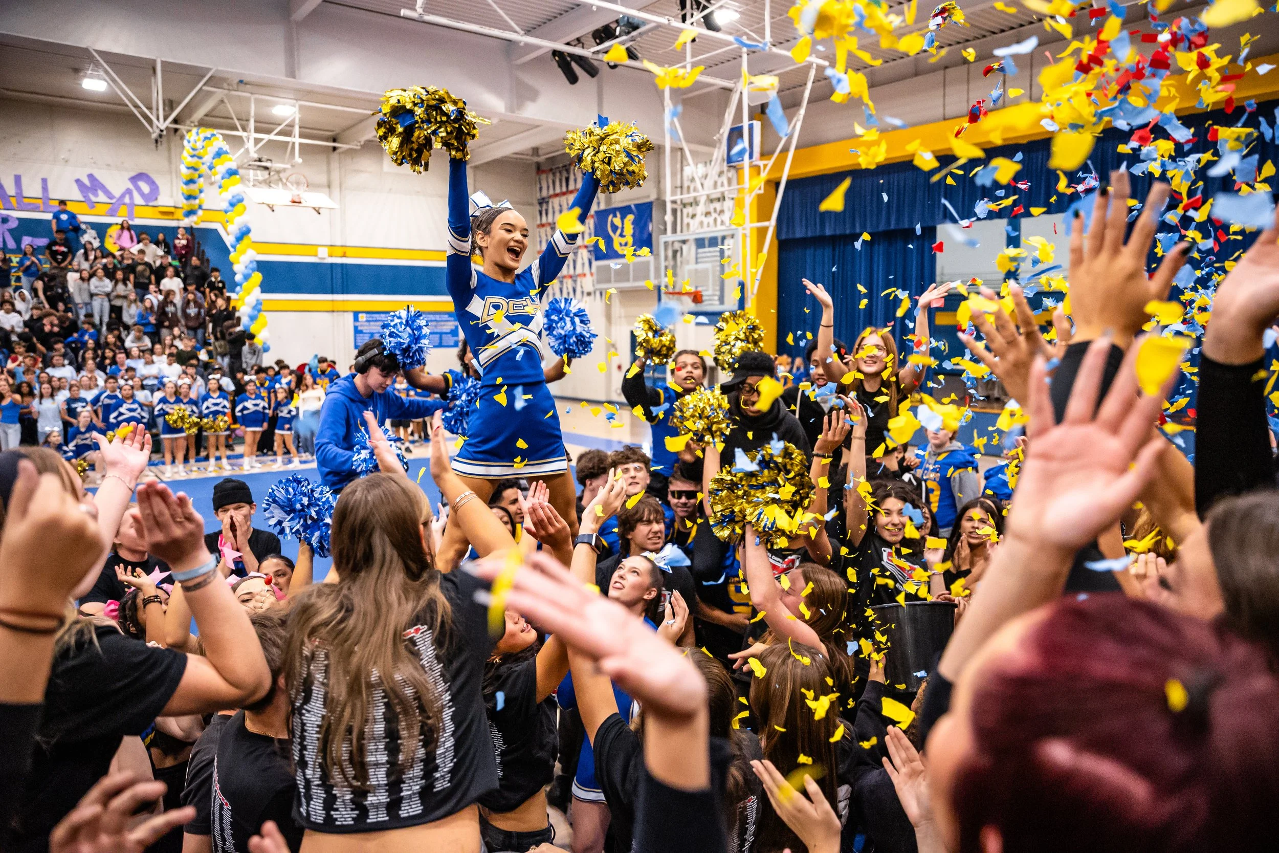 Cheerleaders celebrating at a school gymnasium with confetti falling, surrounded by cheering crowd in the background.