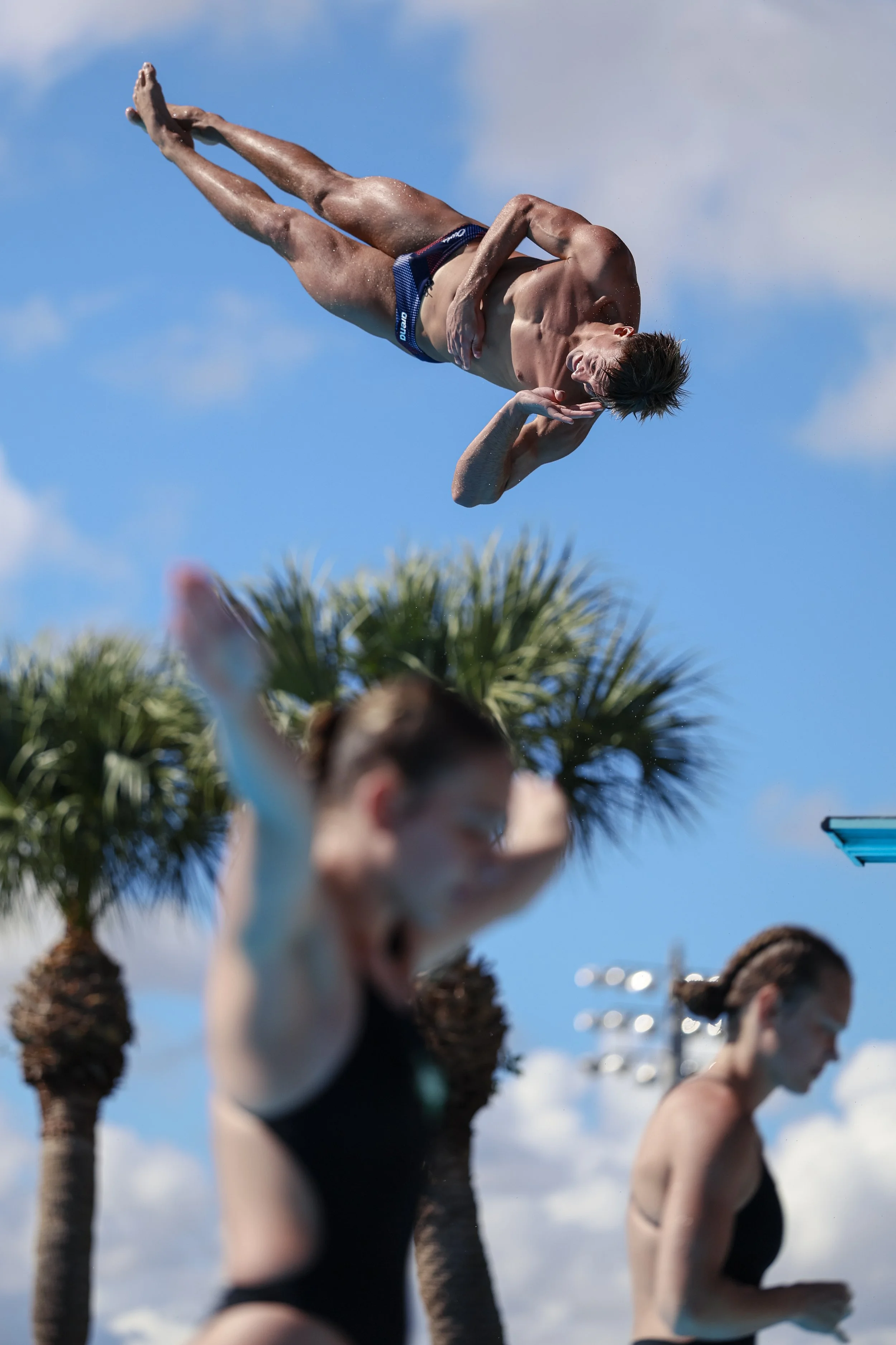 A group of people at the beach wearing swimsuits, with palm trees and a blue sky in the background.