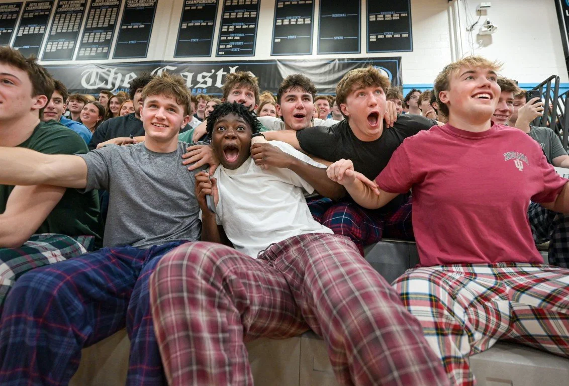 Group of young people sitting in bleachers at a sports event, wearing plaid pajama pants, showing excitement and surprise.