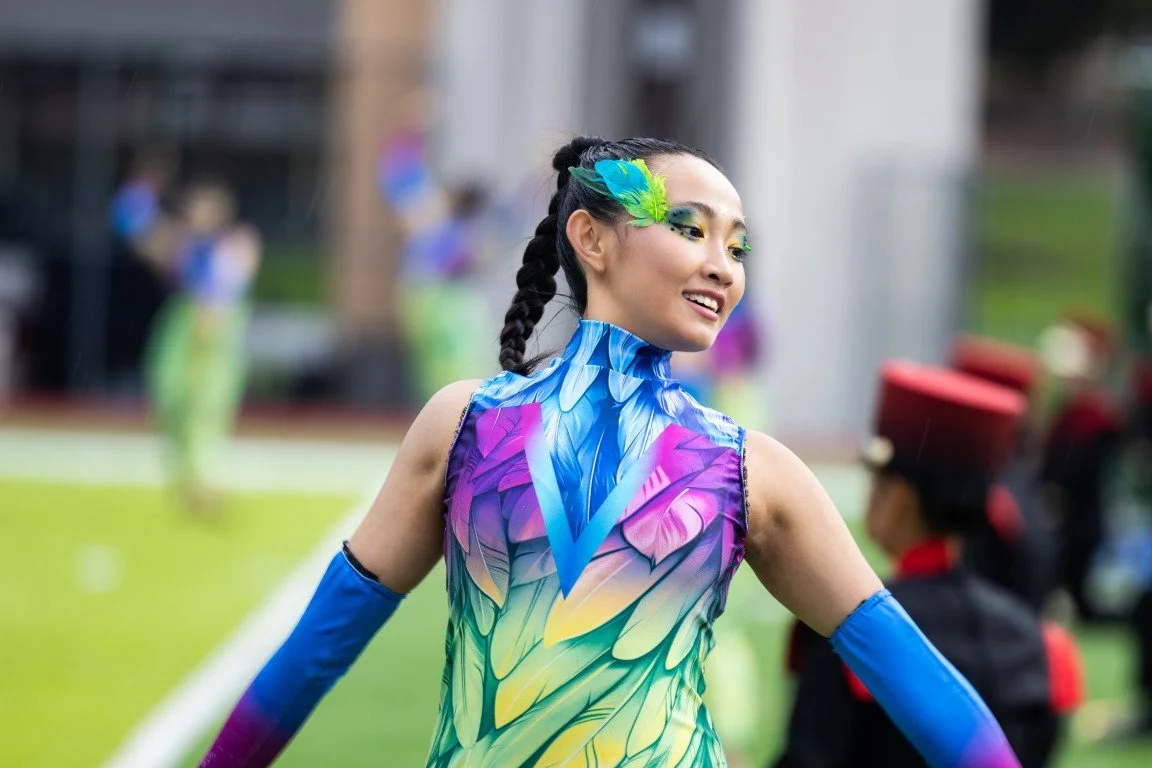 A young woman in a colorful, feather-themed costume with green and purple accents, smiling and looking to the side at an outdoor event.