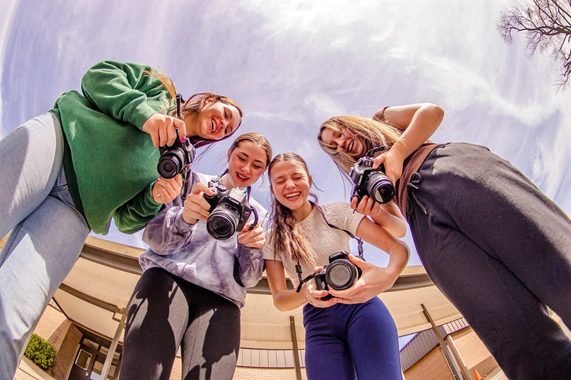 Four young women are standing outdoors, holding cameras, and looking down at the camera, smiling. The photo is taken from a low angle, capturing the clear sky and a building in the background.